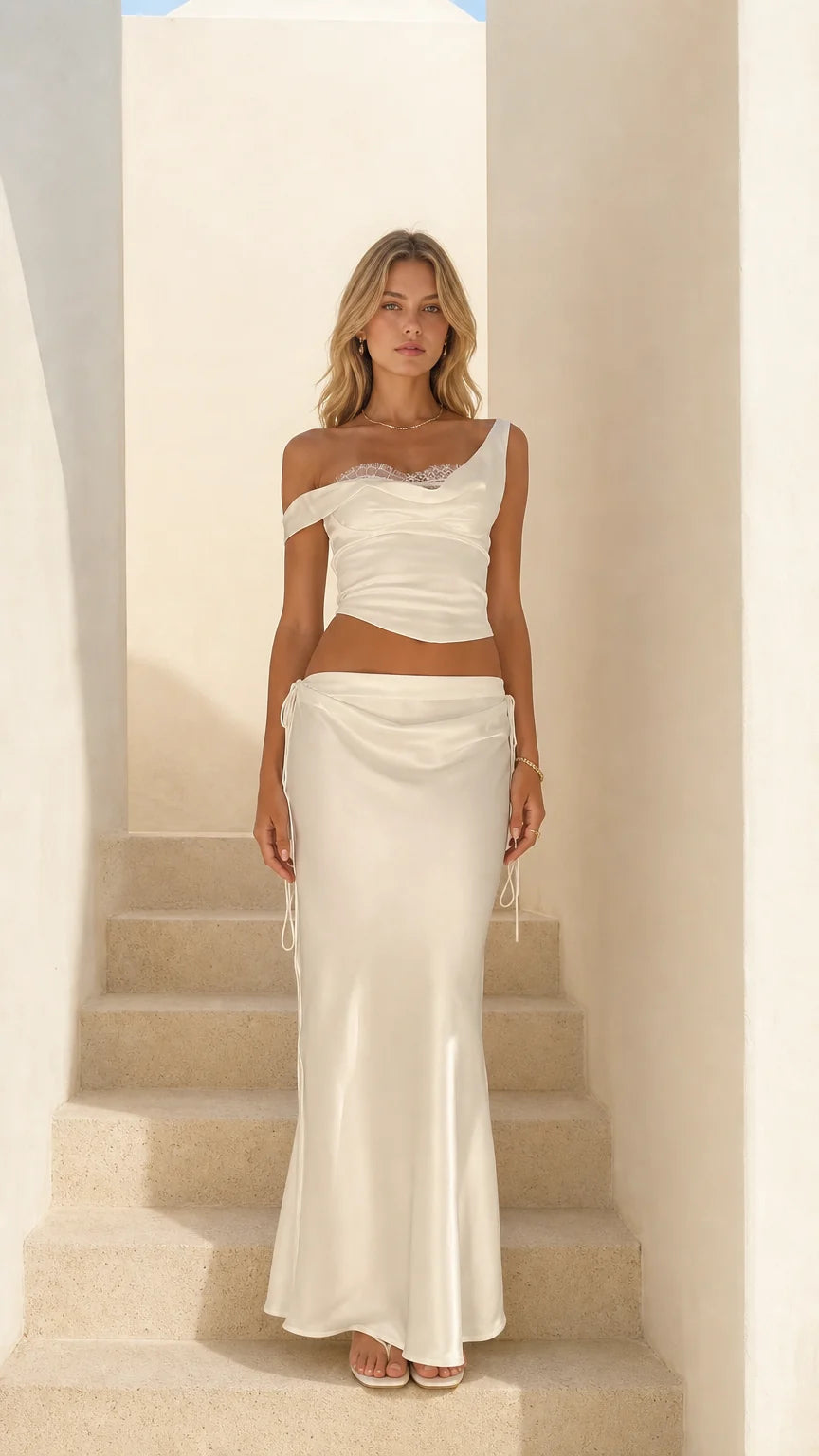 Front view of woman wearing white satin lace-trim crop top and midi skirt set on stairs
